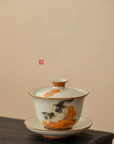 Porcelain gaiwan (lidded bowl) with hand-painted cat and persimmon design, placed on a wooden surface against a beige background