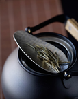Hammered stainless steel tea scoop (8.3 × 3.5 cm) resting on the lid of a black teapot, holding loose tea leaves.
