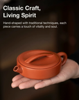 Hand-shaped terracotta teapot held in a hand with text about traditional techniques and vitality.
