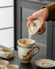 Person pouring tea from a porcelain gaiwan (lidded bowl) into a fairness pitcher, both decorated with hand-painted cat and persimmon design, on a wooden tea table