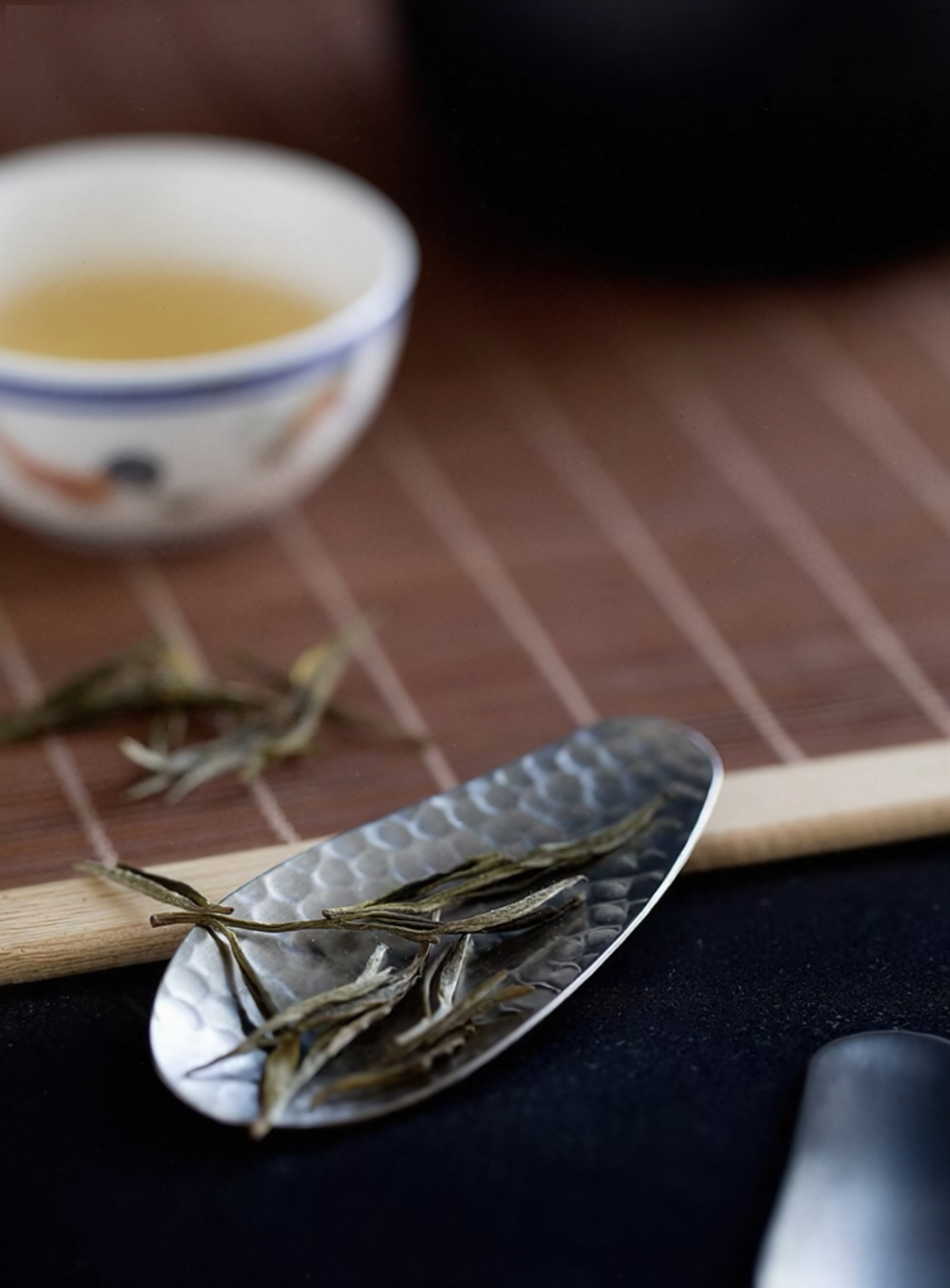Hammered stainless steel tea scoop (8.3 × 3.5 cm) resting on the lid of a black teapot, holding loose tea leaves.