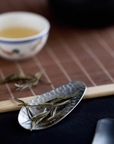 Hammered stainless steel tea scoop (8.3 × 3.5 cm) resting on the lid of a black teapot, holding loose tea leaves.