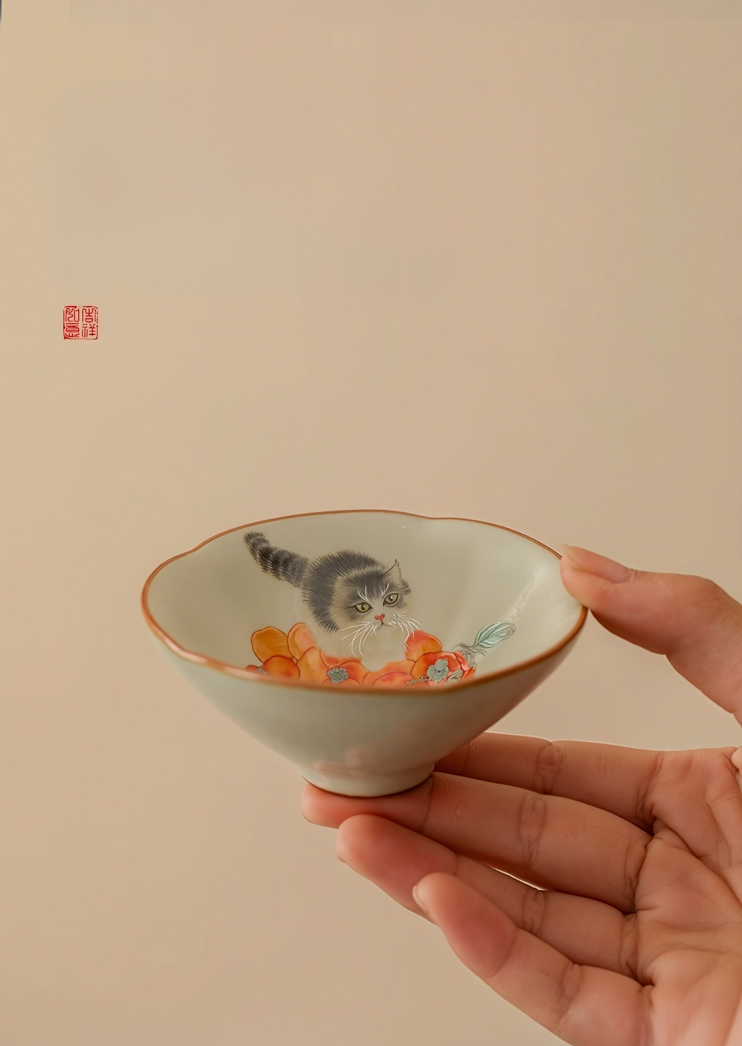 Hand holding a hand-painted ceramic douli teacup with kitten and persimmon design against a beige background
