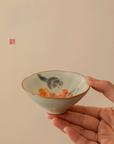 Hand holding a hand-painted ceramic douli teacup with kitten and persimmon design against a beige background
