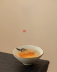 Hand-painted ceramic douli teacup with kitten and persimmon design, filled with tea, placed on a wooden surface against a beige background