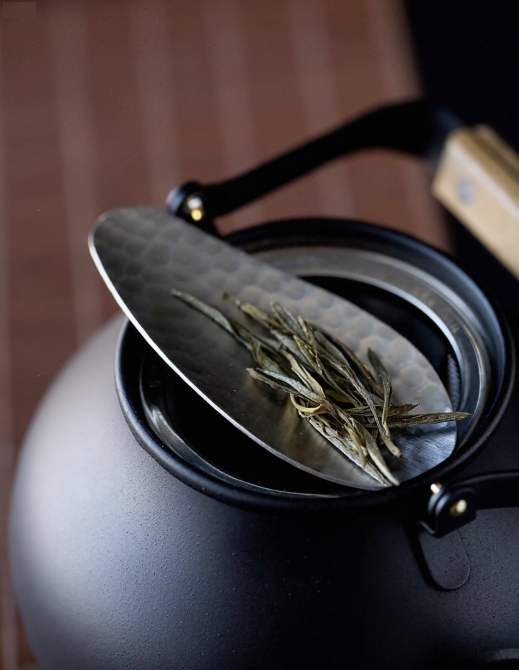 Hammered stainless steel tea scoop (8.3 × 3.5 cm) resting on the lid of a black teapot, holding loose tea leaves.
