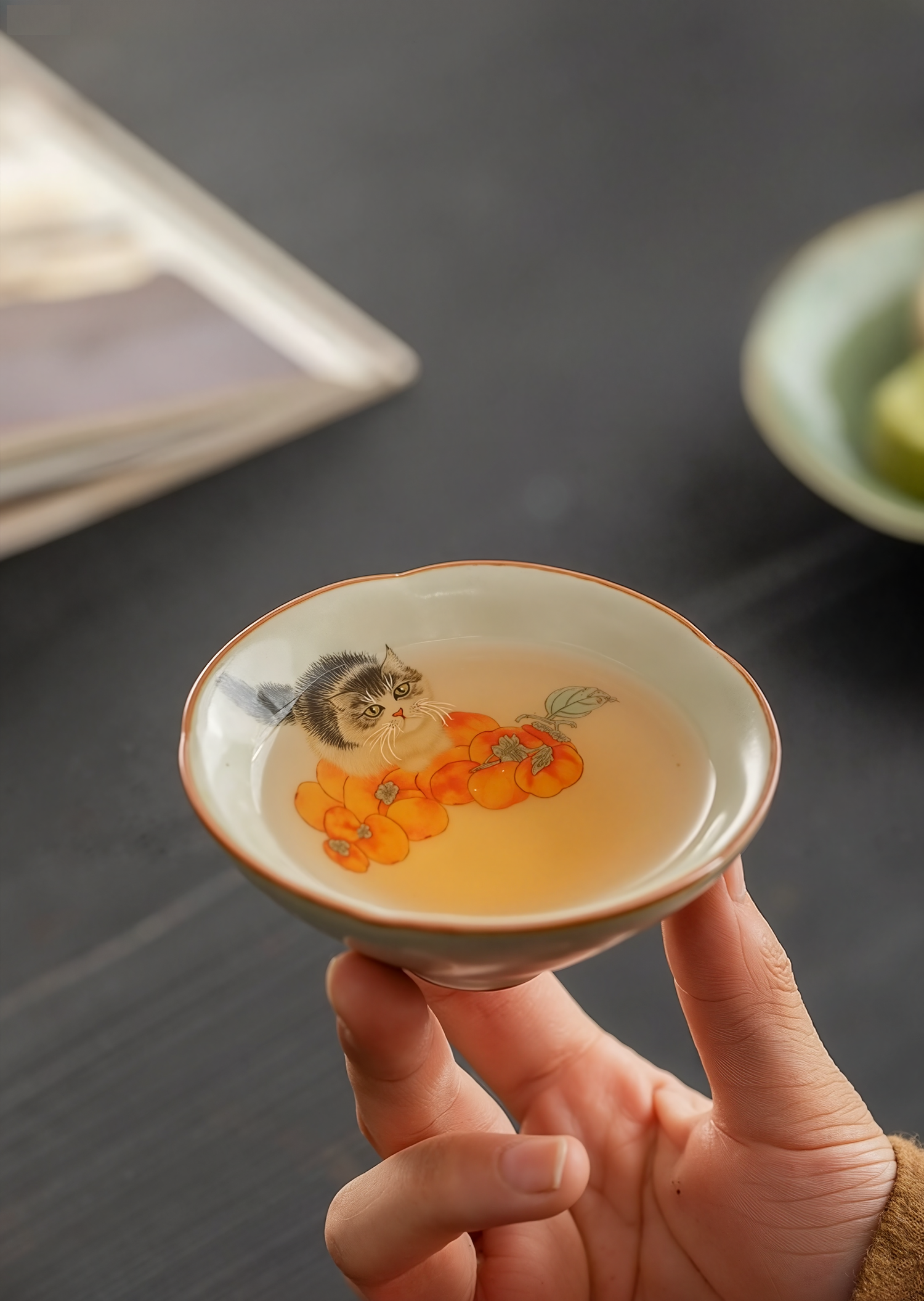Hand holding a douli teacup with kitten and persimmon design, filled with tea, against a dark tabletop background