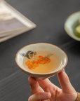Hand holding a douli teacup with kitten and persimmon design, filled with tea, against a dark tabletop background