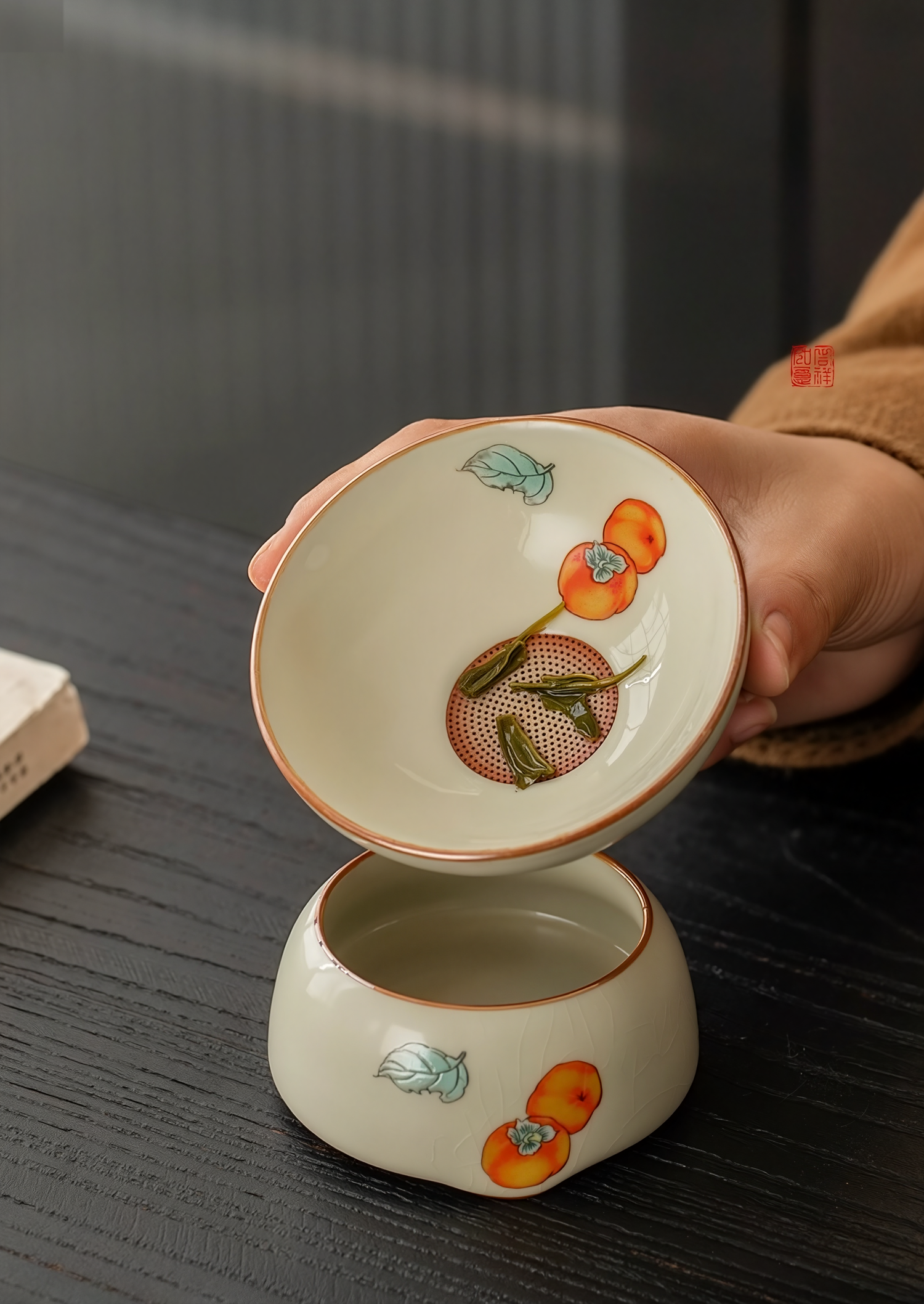 Porcelain tea strainer with hand-painted persimmon design, shown in use on a wooden table