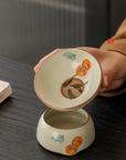 Porcelain tea strainer with hand-painted persimmon design, shown in use on a wooden table