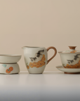 Three-piece ceramic tea set with cat and persimmon design, including gaiwan, fairness pitcher, and tea strainer, on a plain beige background.