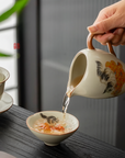 Pouring tea from a hand-painted ceramic fairness pitcher into a douli teacup with kitten and persimmon design on a wooden table