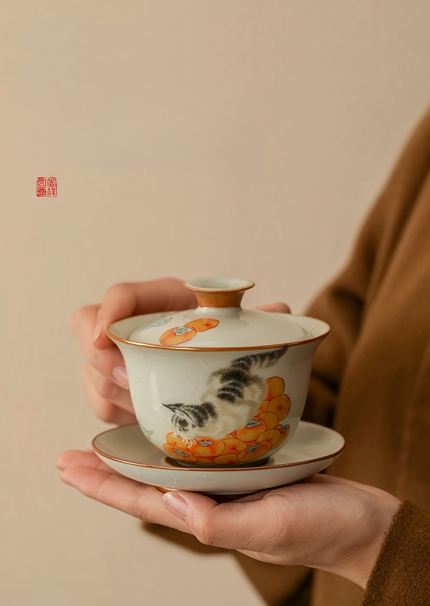 Person holding a porcelain gaiwan (lidded bowl) with hand-painted cat and persimmon design against a beige background