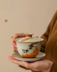 Person holding a porcelain gaiwan (lidded bowl) with hand-painted cat and persimmon design against a beige background
