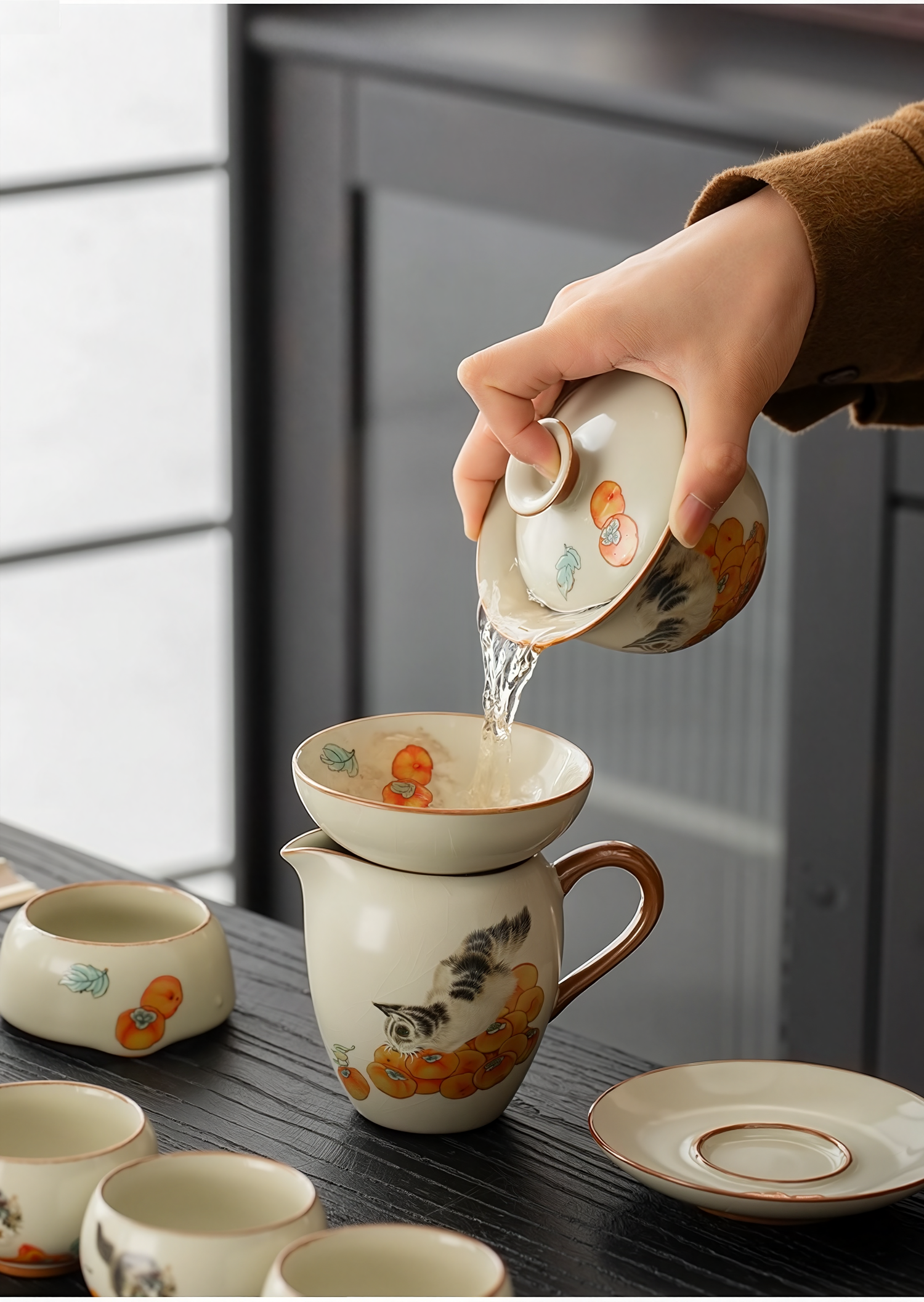 Person pouring tea from a porcelain gaiwan (lidded bowl) into a fairness pitcher, both decorated with hand-painted cat and persimmon design, on a wooden tea table