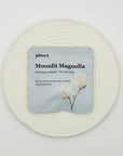 Moonlit Magnolia DanCong Oolong tea bag from Jaktea placed on a white textured plate, featuring soft magnolia blossom artwork on a pale blue pouch.