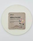 Silken Orchid DanCong Oolong tea bag by Jaktea, displayed on a textured white plate with soft floral packaging.