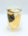 Side view of Silken Orchid Dancong Oolong cold brew tea, with the pyramid tea bag floating among ice inside a clear double-wall glass.