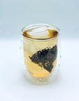 Cold-brew Sunlit Petal DanCong Oolong tea bag steeping in a clear double-wall glass, showing floral leaves unfolding in pale golden tea.